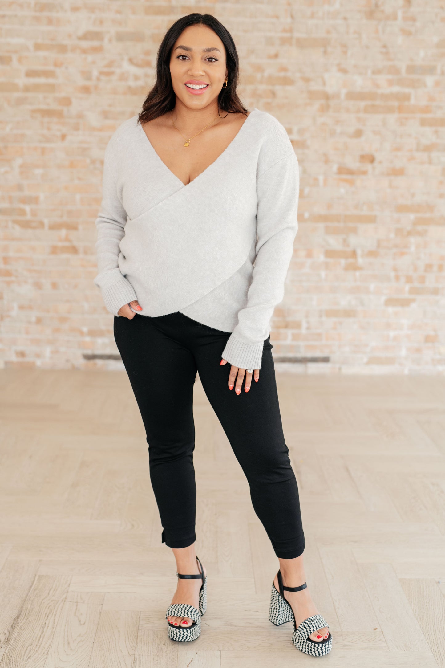 Woman wearing a light gray sweater and black pants standing against a brick wall.