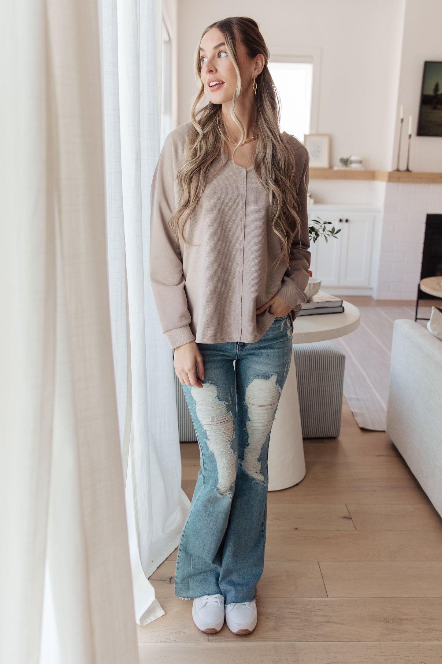 Woman standing in a modern living room wearing a beige sweater and blue jeans.