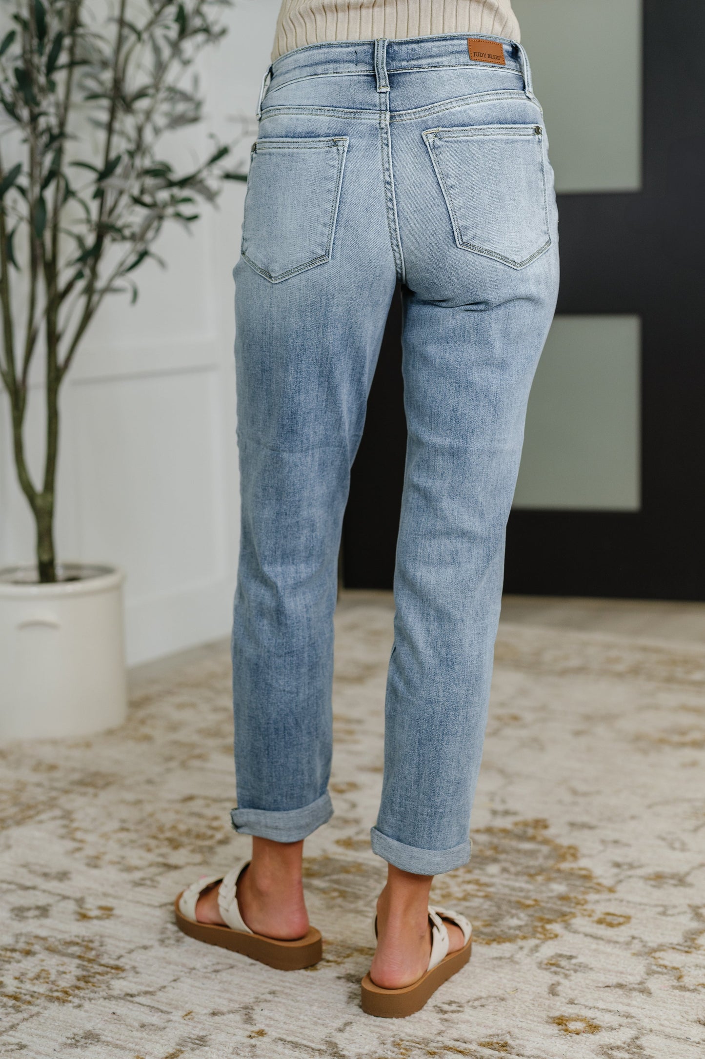 Person wearing light blue jeans standing in a room with a plant and rug.