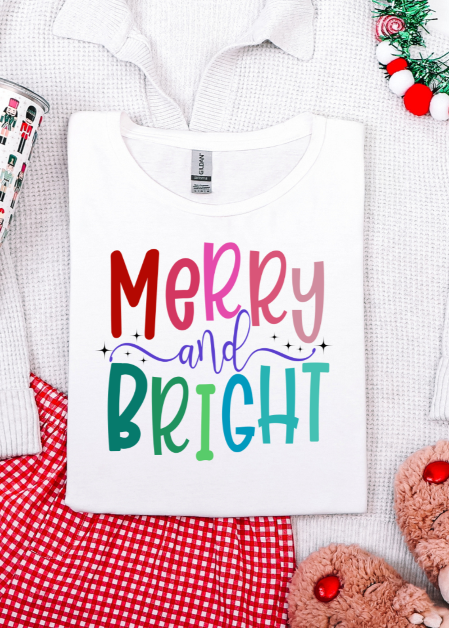 White t-shirt with 'Merry and Bright' text, surrounded by Christmas-themed items on a white background.