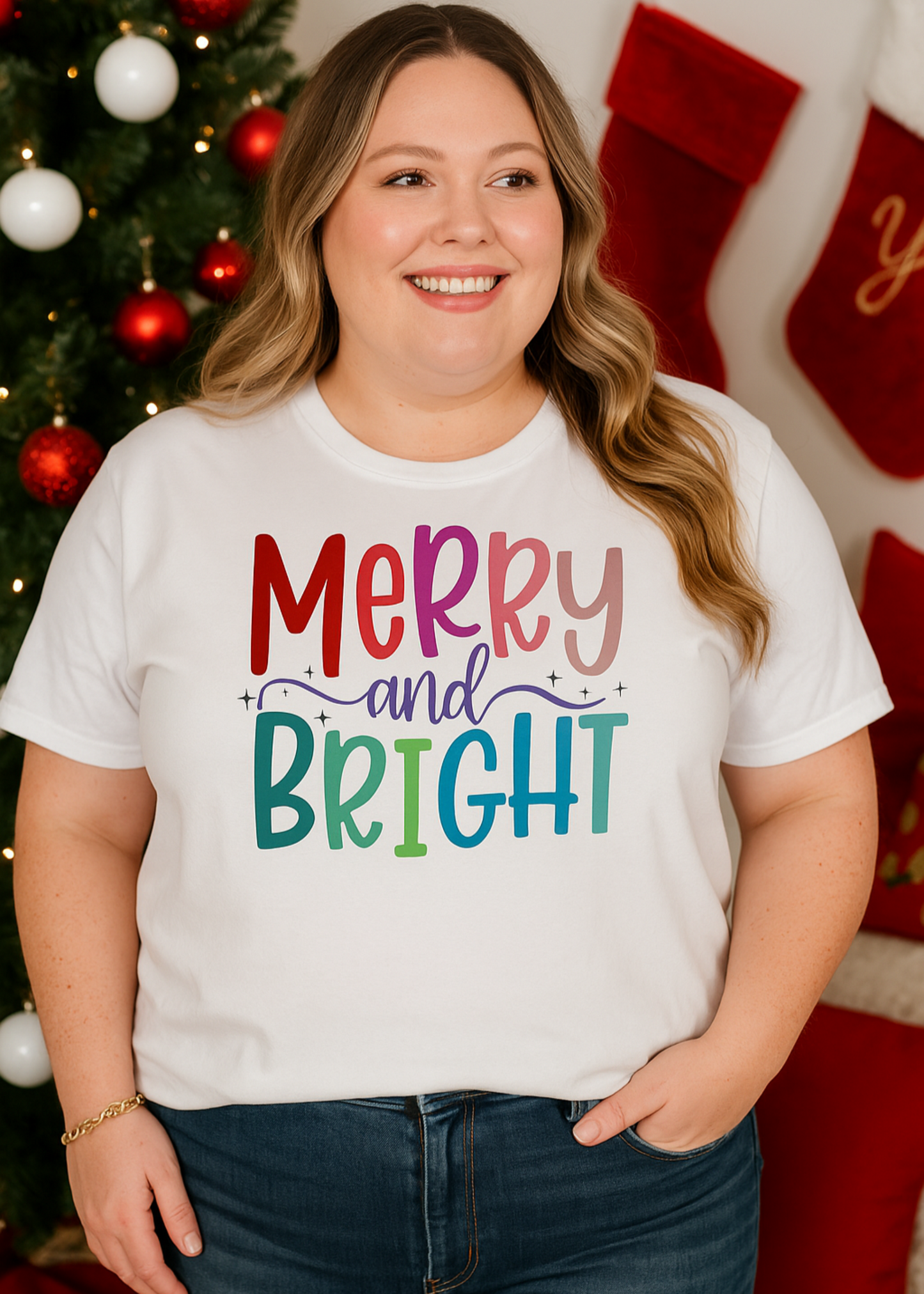Person wearing a white t-shirt with 'Merry and Bright' text in colorful letters, standing in front of a decorated Christmas tree and stockings.