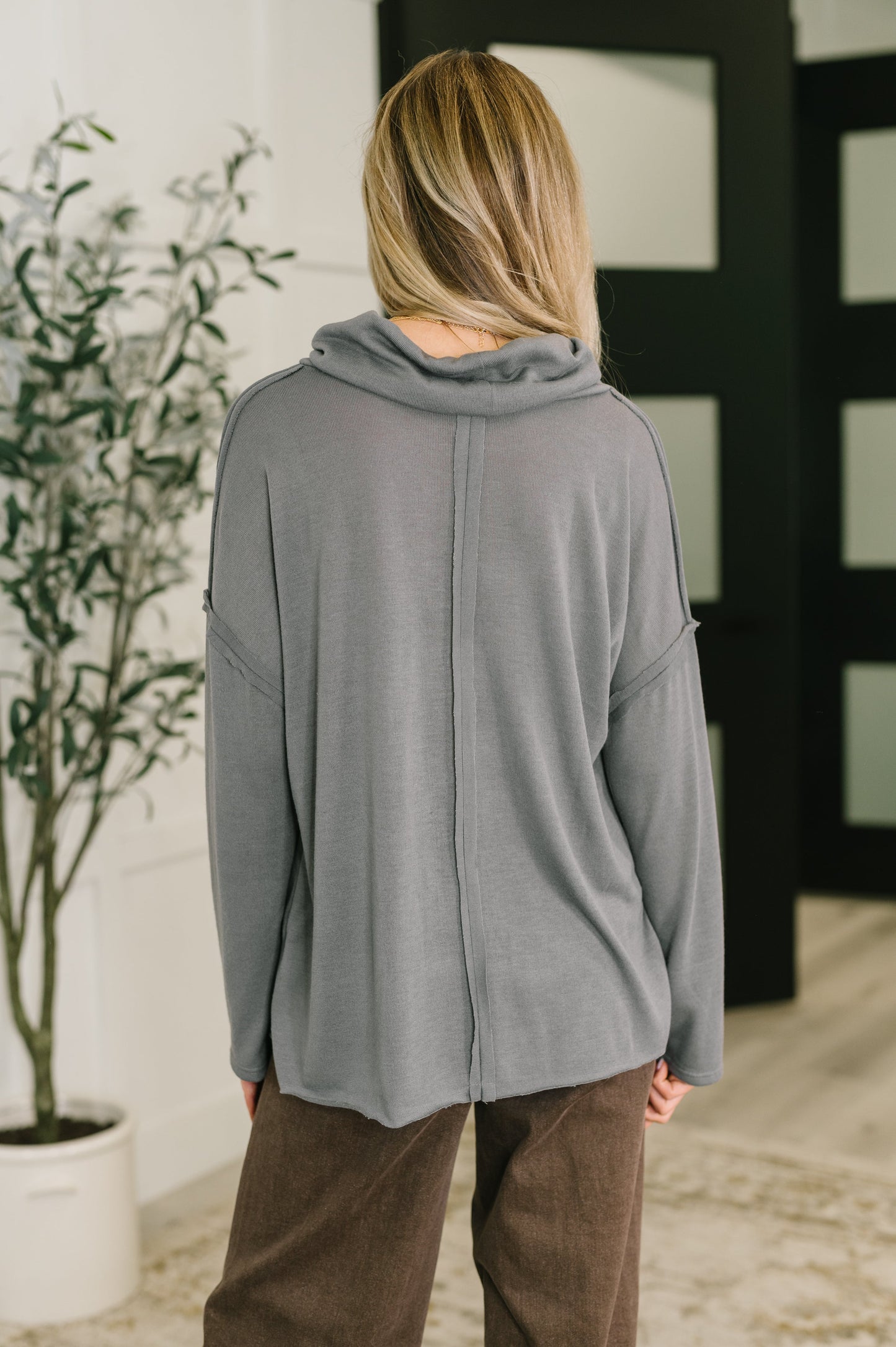 Person wearing a gray cardigan and brown pants standing in a room with a plant and decorative wall.
