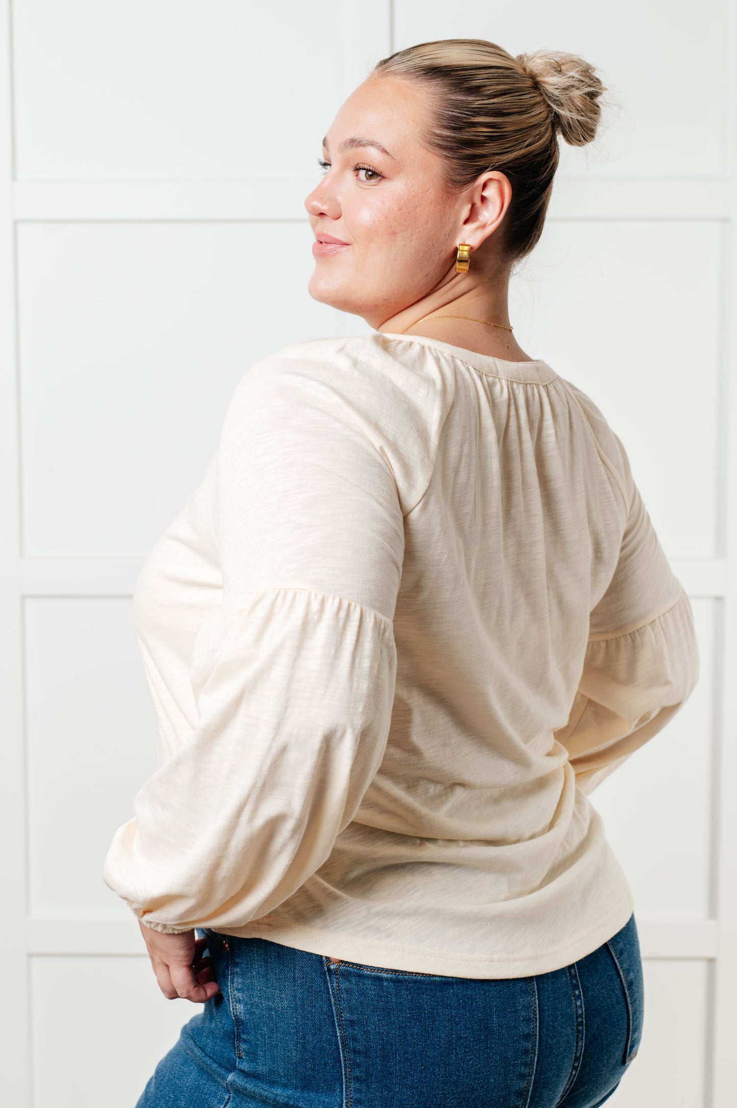 Woman wearing a beige blouse with puffed sleeves and blue jeans against a white tiled wall.