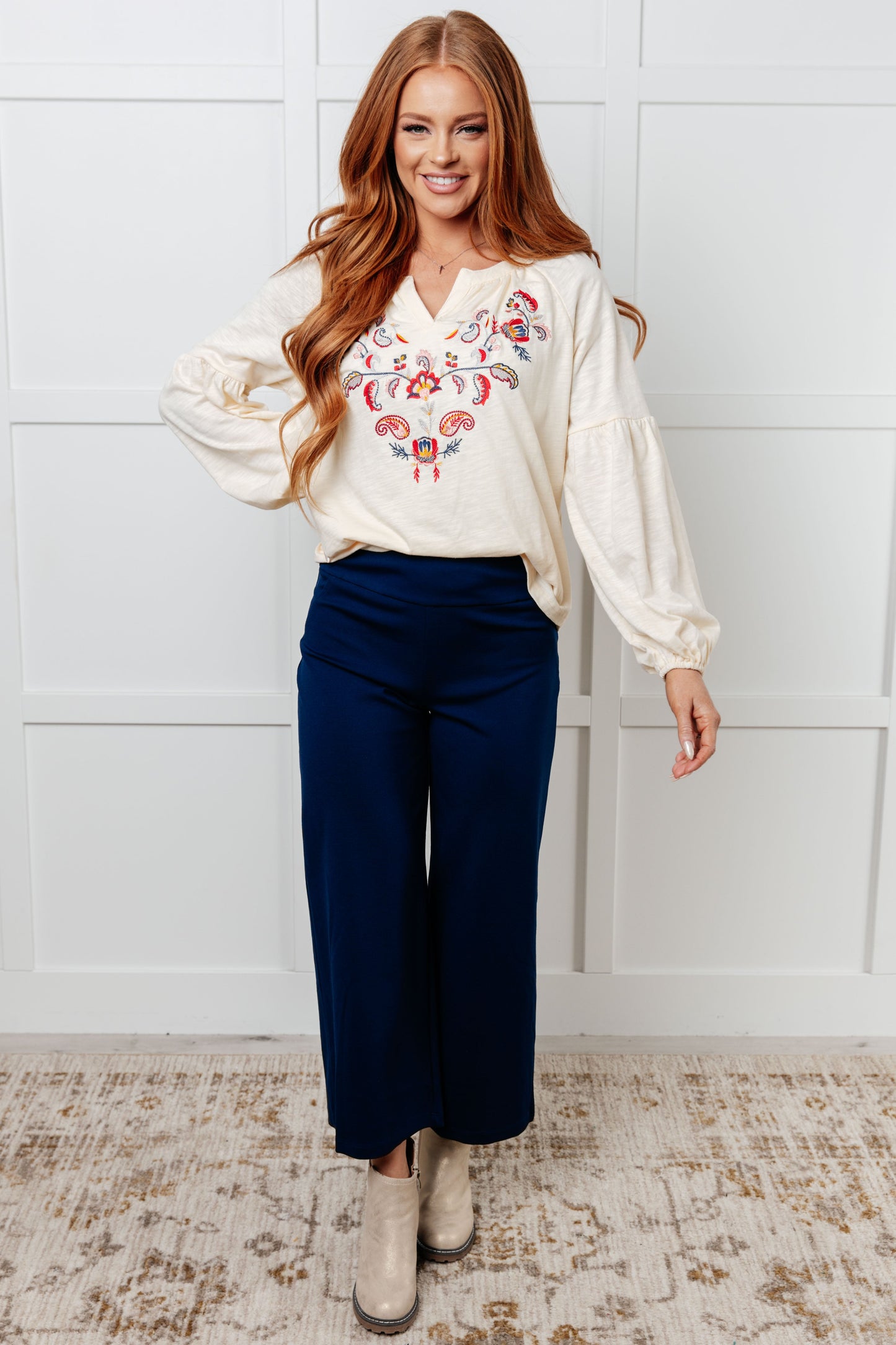 Woman wearing a white blouse with floral embroidery and blue pants against a white wall.