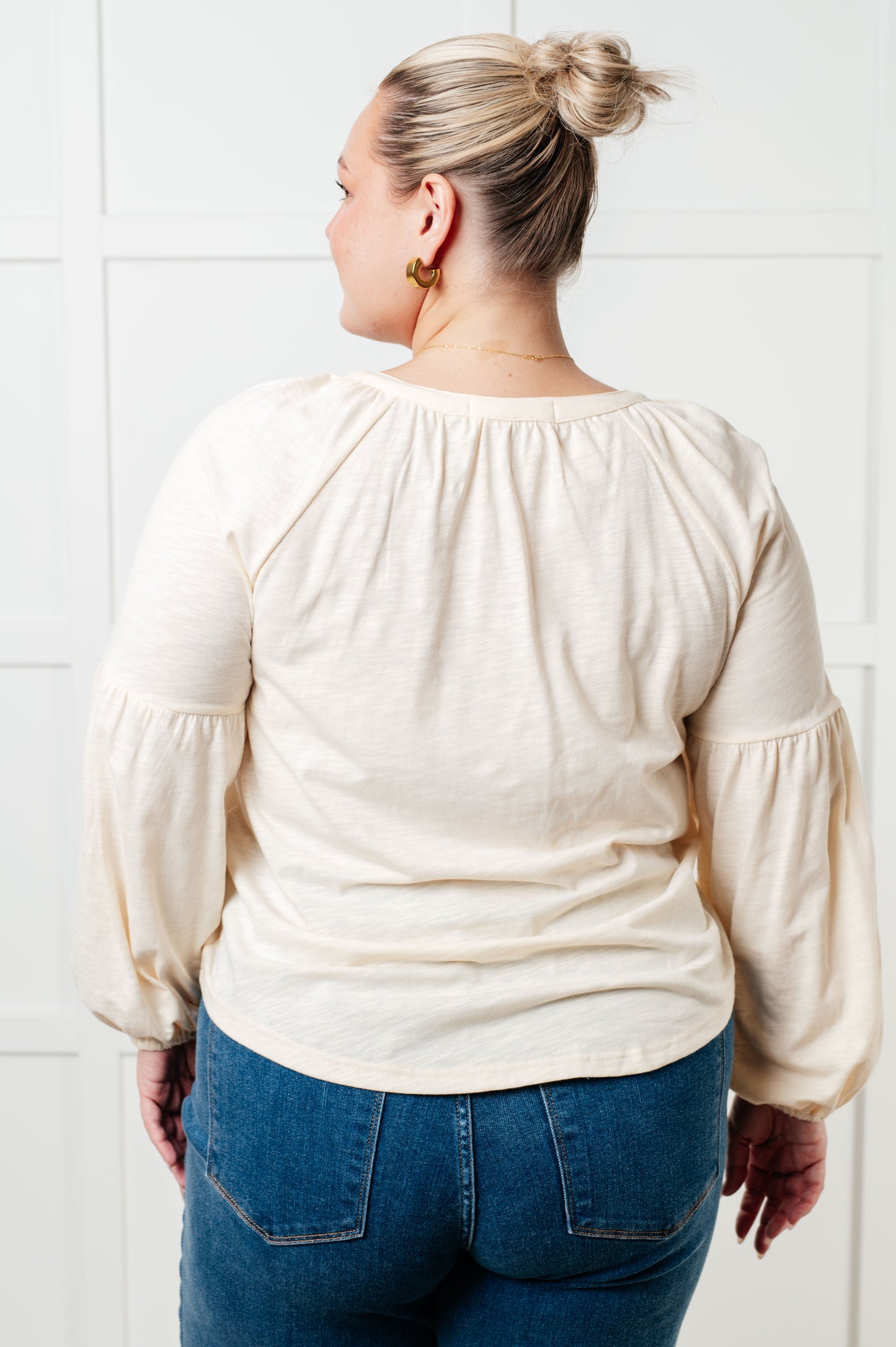Person wearing a cream blouse with long sleeves and blue jeans against a white tiled wall.