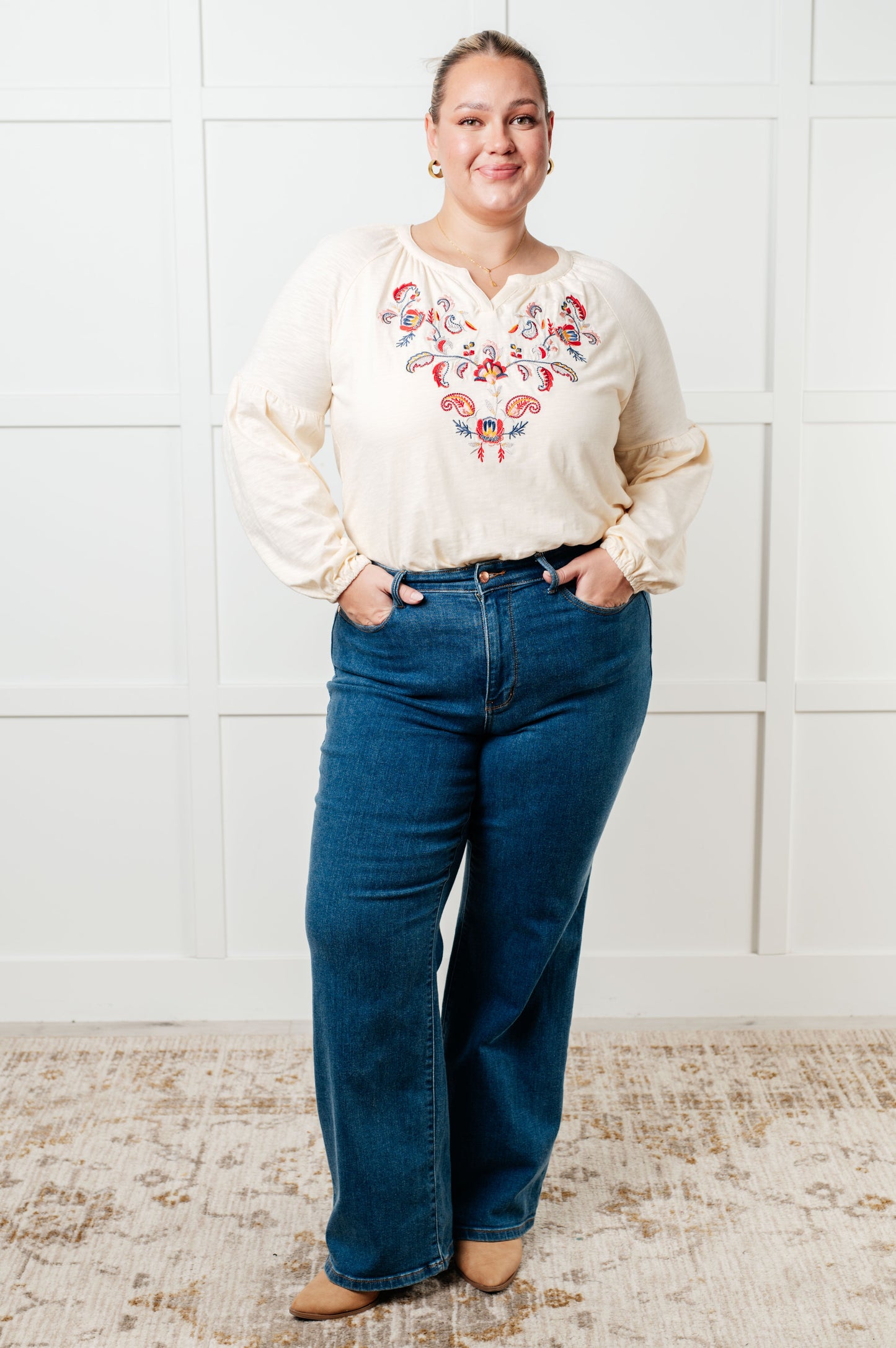Woman wearing a white blouse with floral embroidery and blue jeans against a white paneled wall.