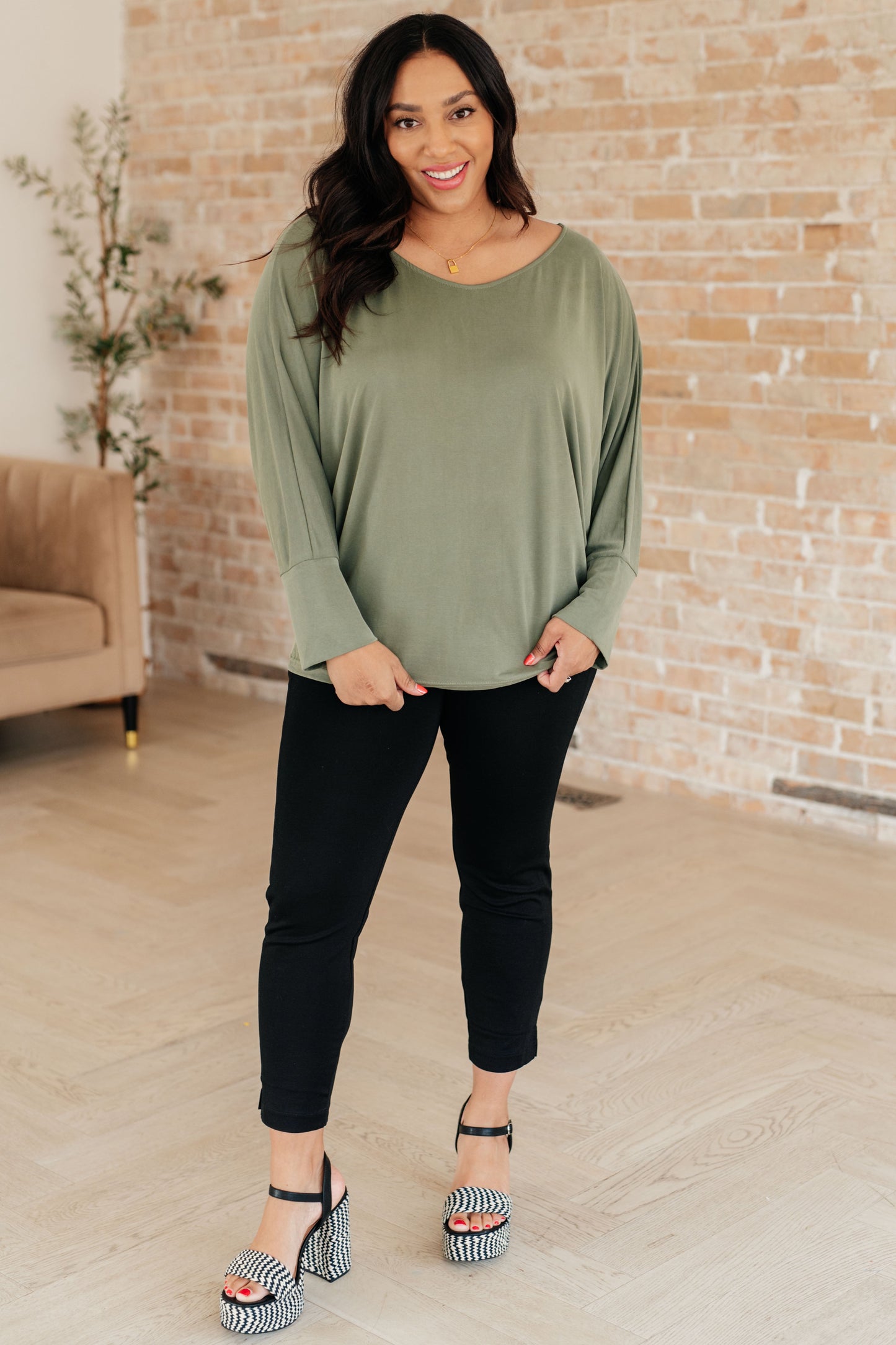 Woman wearing an olive green top and black pants standing in a room with a brick wall.