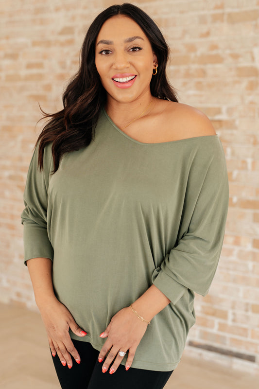 Woman wearing an olive green off-shoulder top against a brick wall.