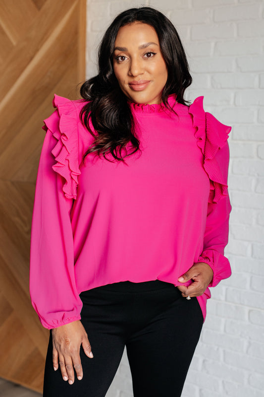 Woman wearing a bright pink blouse with ruffled sleeves against a white brick wall.