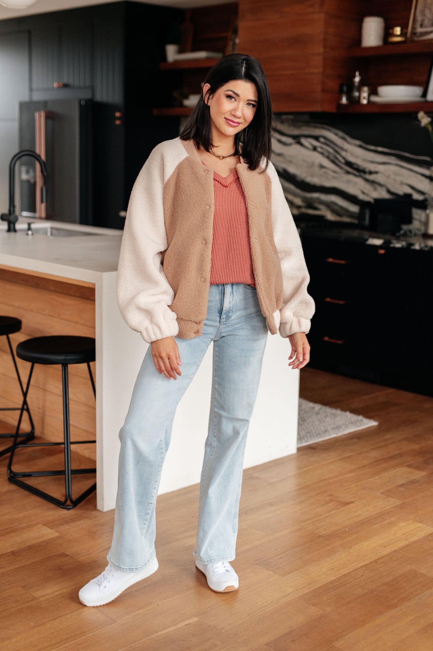 Woman standing in a kitchen wearing a beige and white jacket, pink top, and light blue jeans.