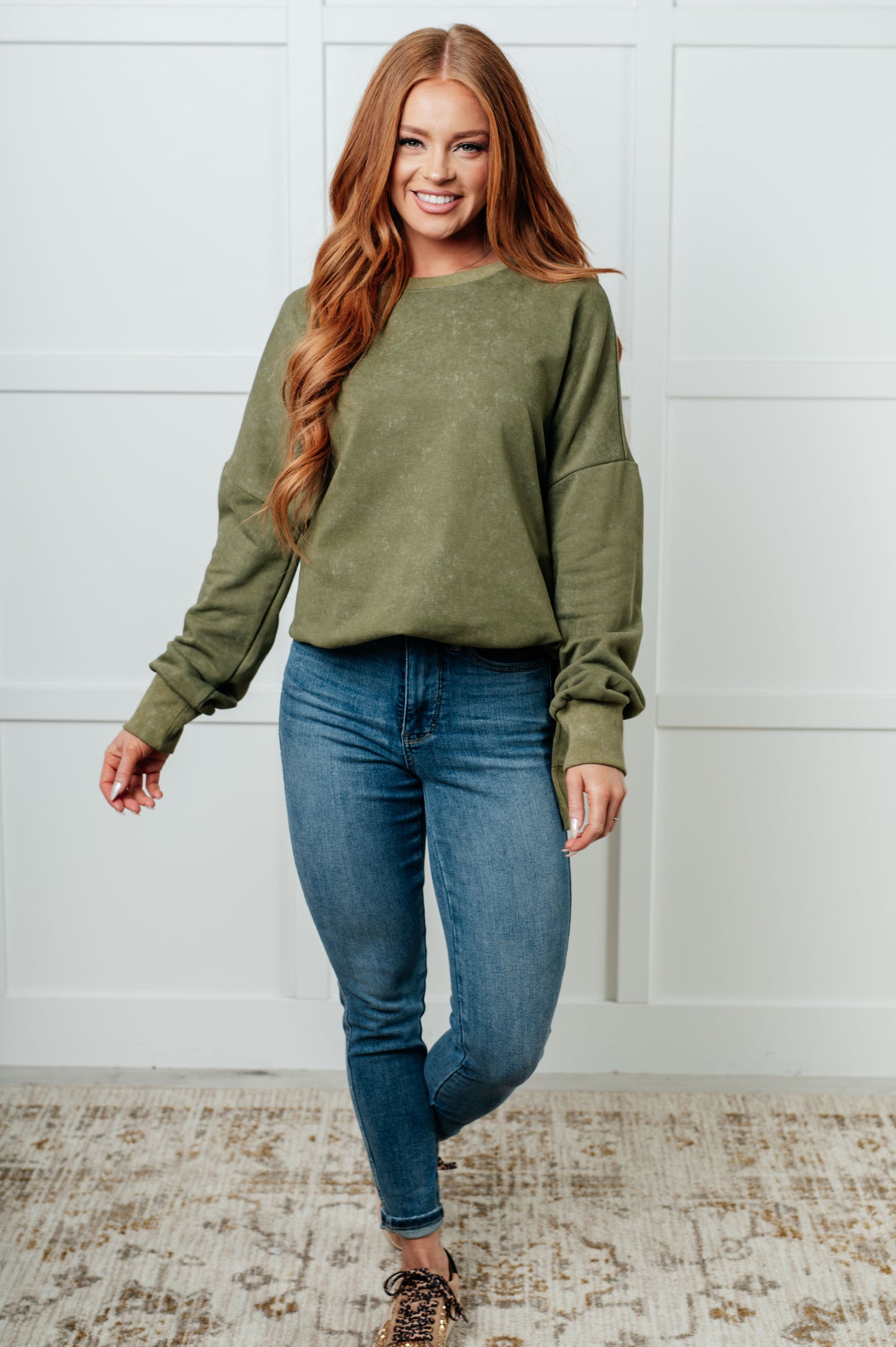 Woman wearing an olive green sweatshirt and blue jeans standing against a white wall.