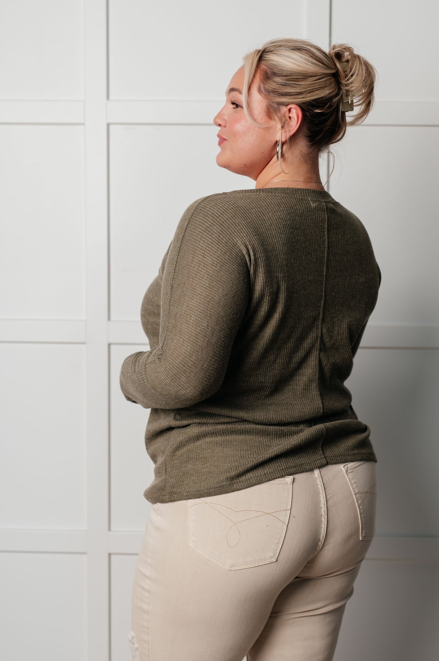 Woman wearing a green sweater and beige pants against a white tiled wall.