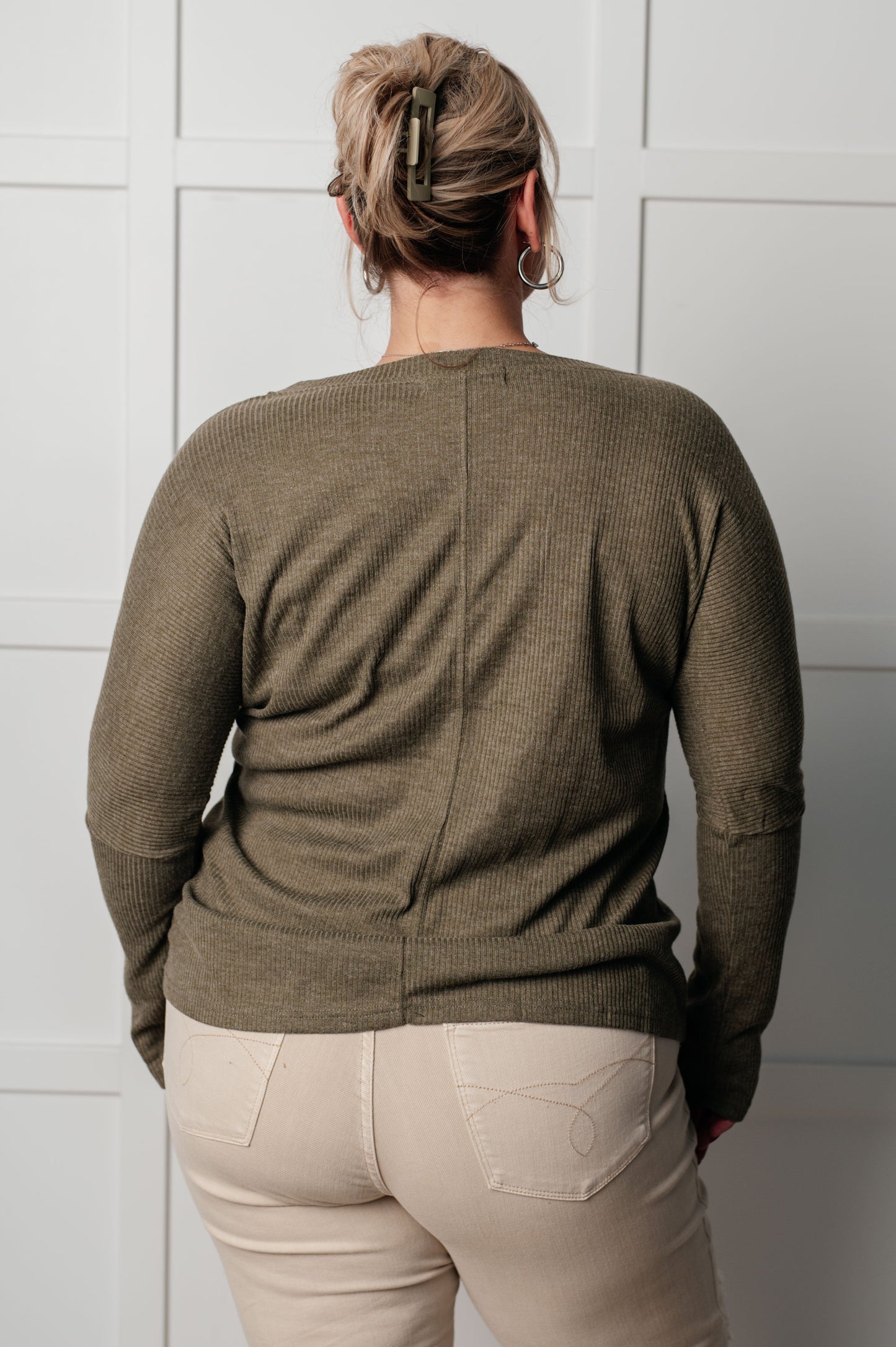 Person wearing a green sweater and beige pants against a white paneled wall.