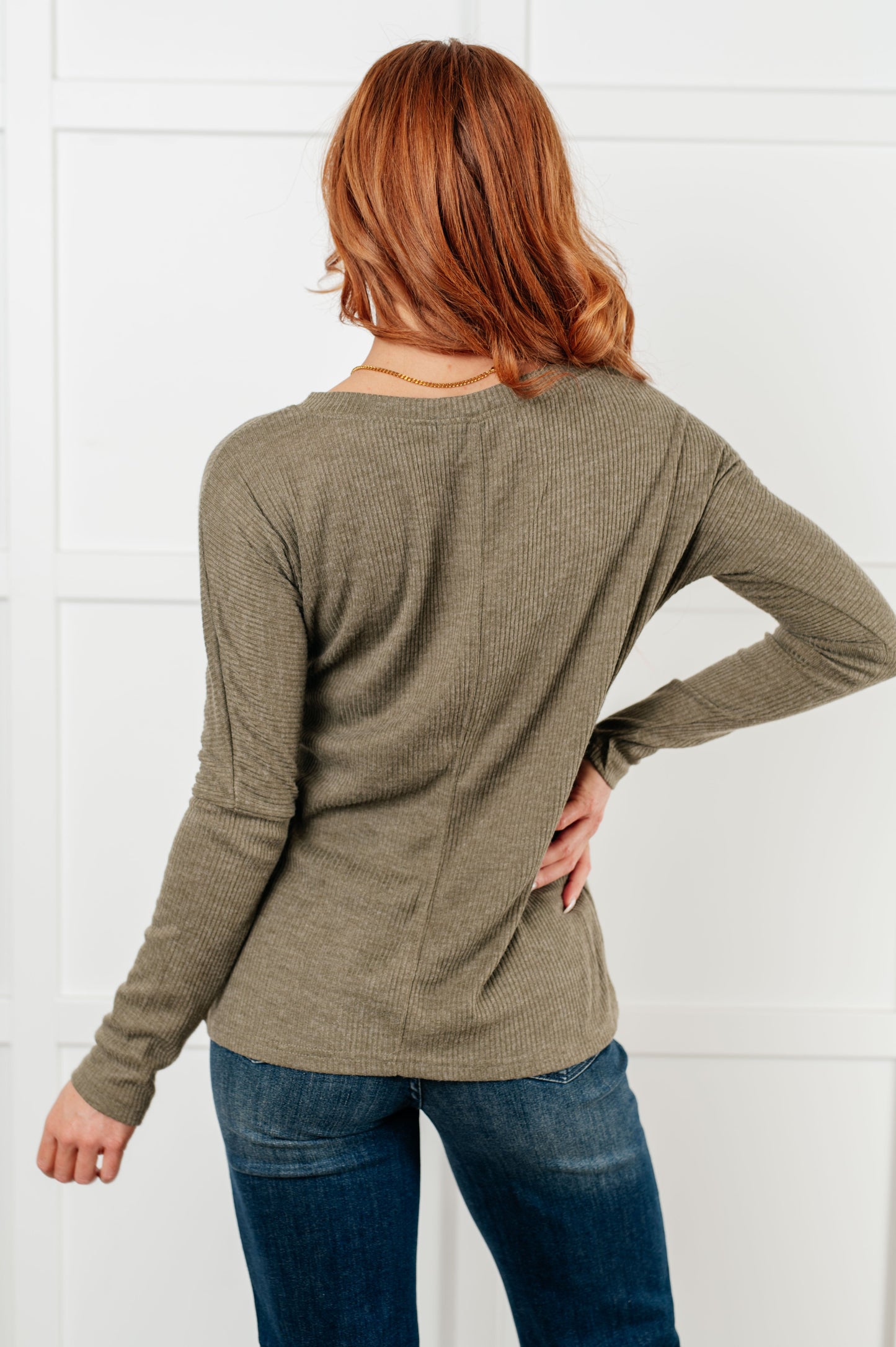 Person wearing a green long-sleeve top and blue jeans against a white tiled wall.