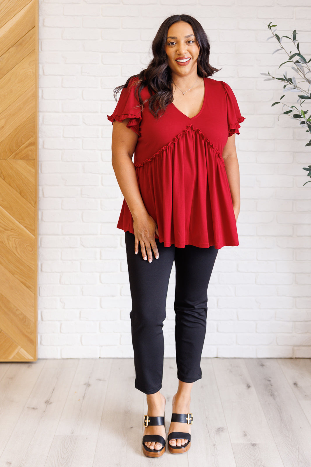 Woman wearing a red blouse and black pants standing in a room with a wooden door and white brick wall.
