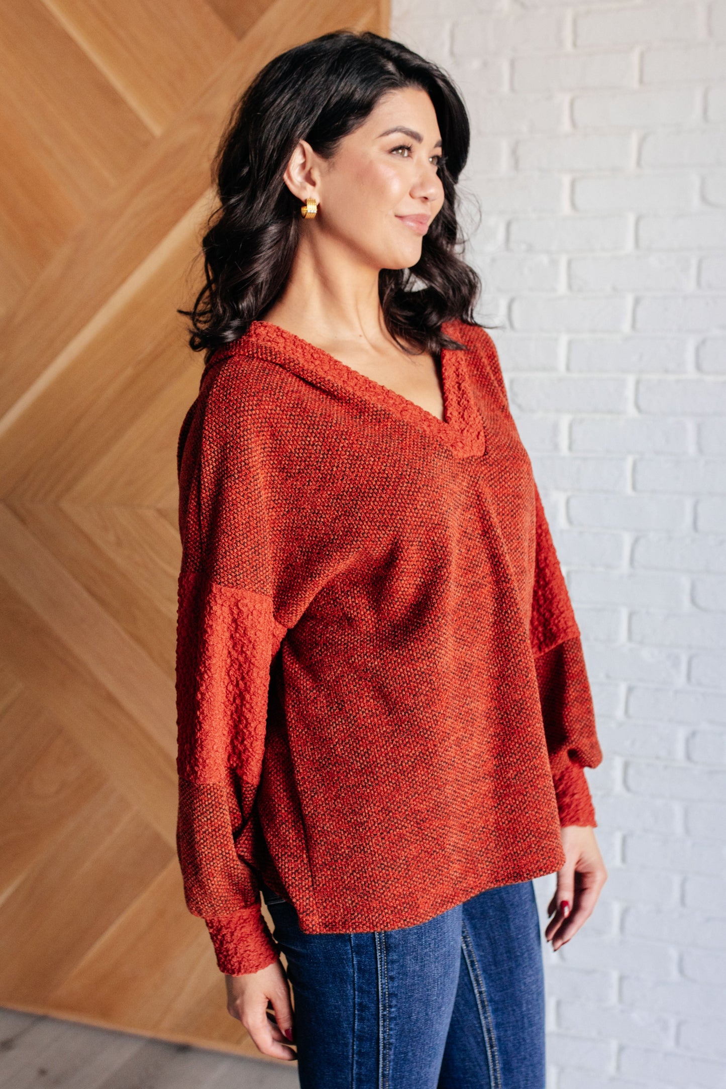 Woman wearing a red sweater and blue jeans against a white brick wall.