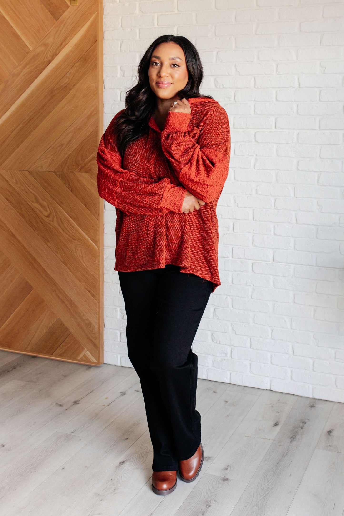 Woman wearing a red sweater and black pants standing against a white brick wall.