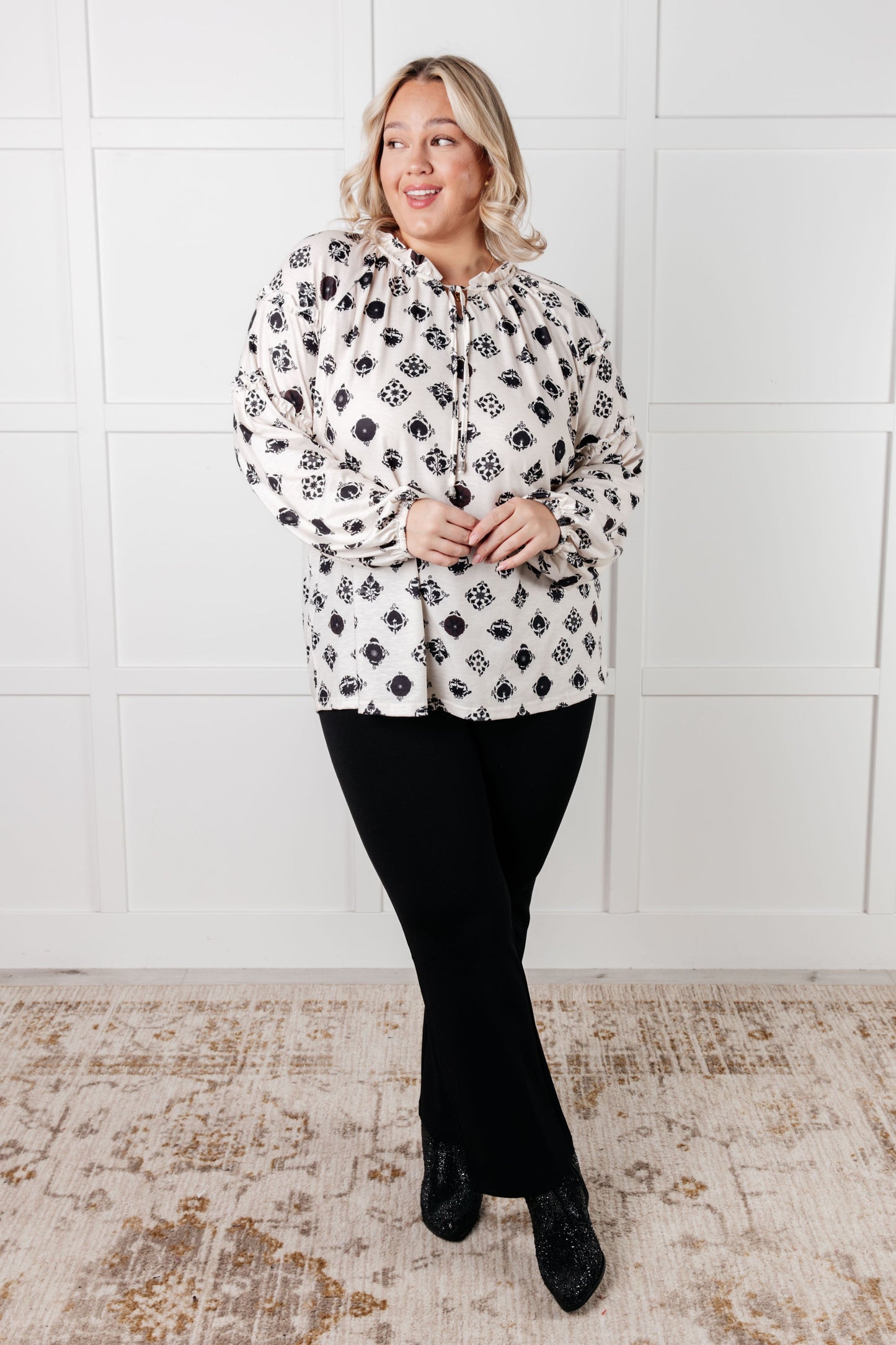Woman wearing a black and white floral blouse with black pants against a white tiled wall.
