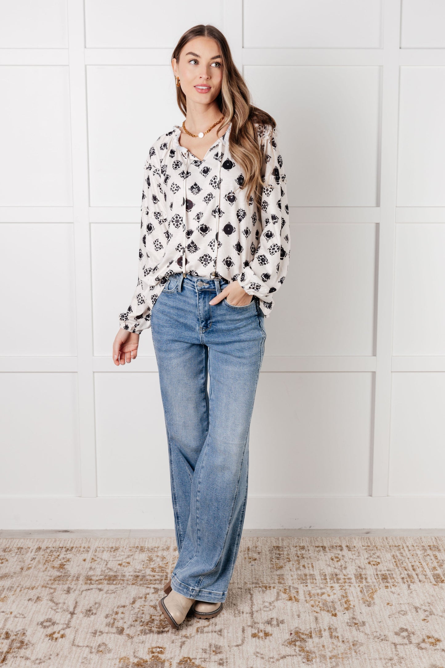 Woman wearing a patterned blouse and jeans against a white tiled wall.