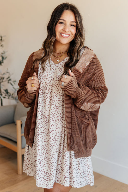 Woman wearing a brown cardigan over a white dress with black patterns, standing indoors.