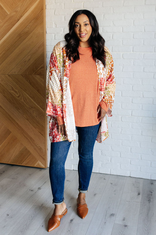 Woman wearing a floral kimono over a pink top and blue jeans, standing in a room with wooden and white brick walls.