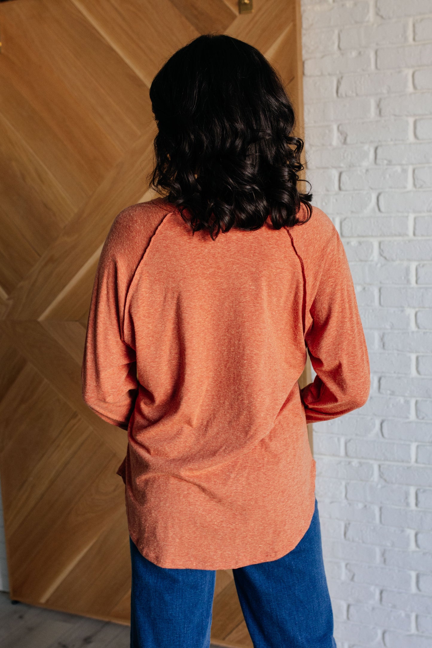 Person wearing an orange sweater and blue pants standing against a wooden and white brick wall.