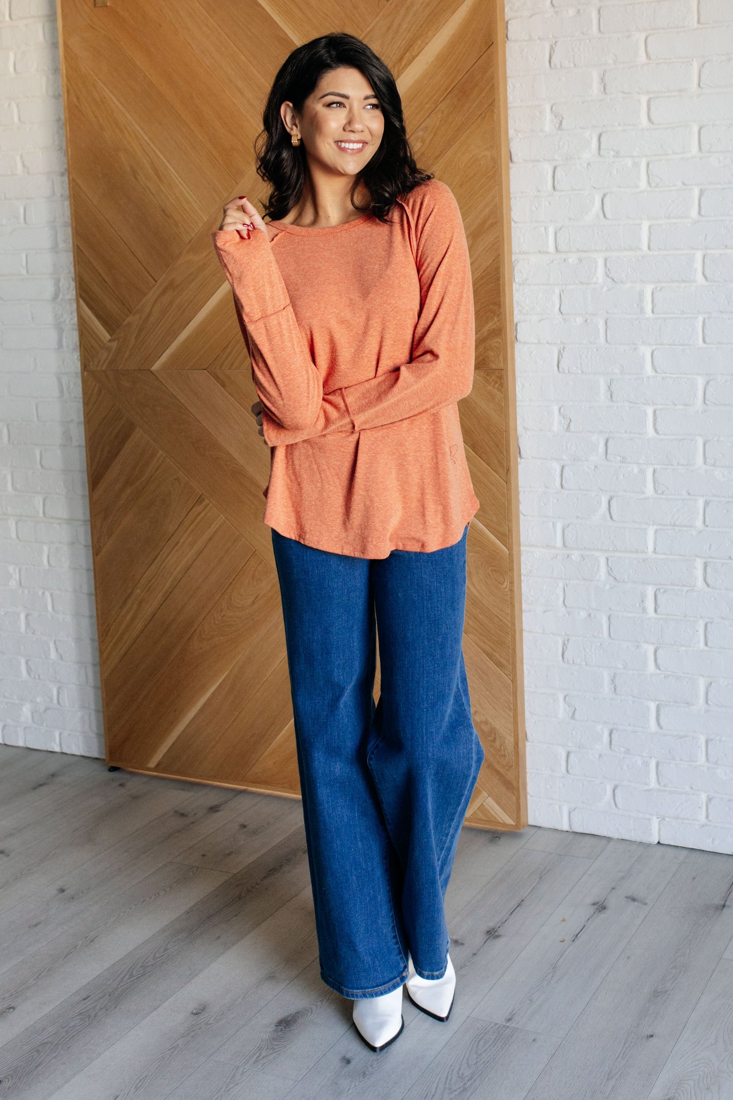 Woman wearing an orange top and blue jeans standing in a room with wooden paneling and white brick wall.