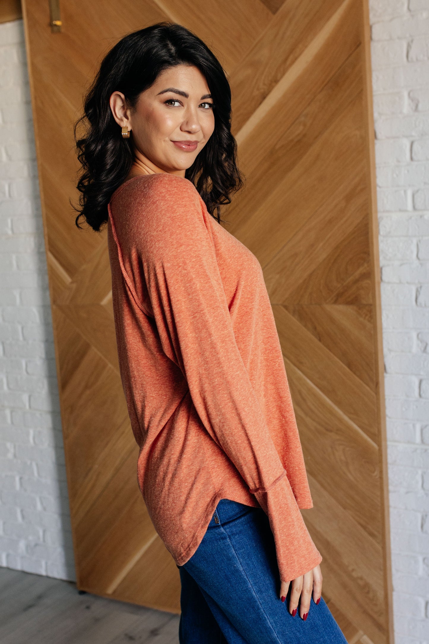 Woman wearing an orange sweater and blue jeans standing against a wooden wall.
