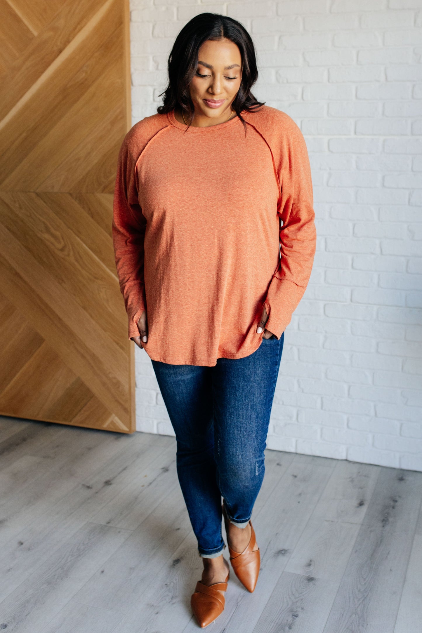 Woman wearing an orange long-sleeve top and blue jeans standing against a white brick wall.