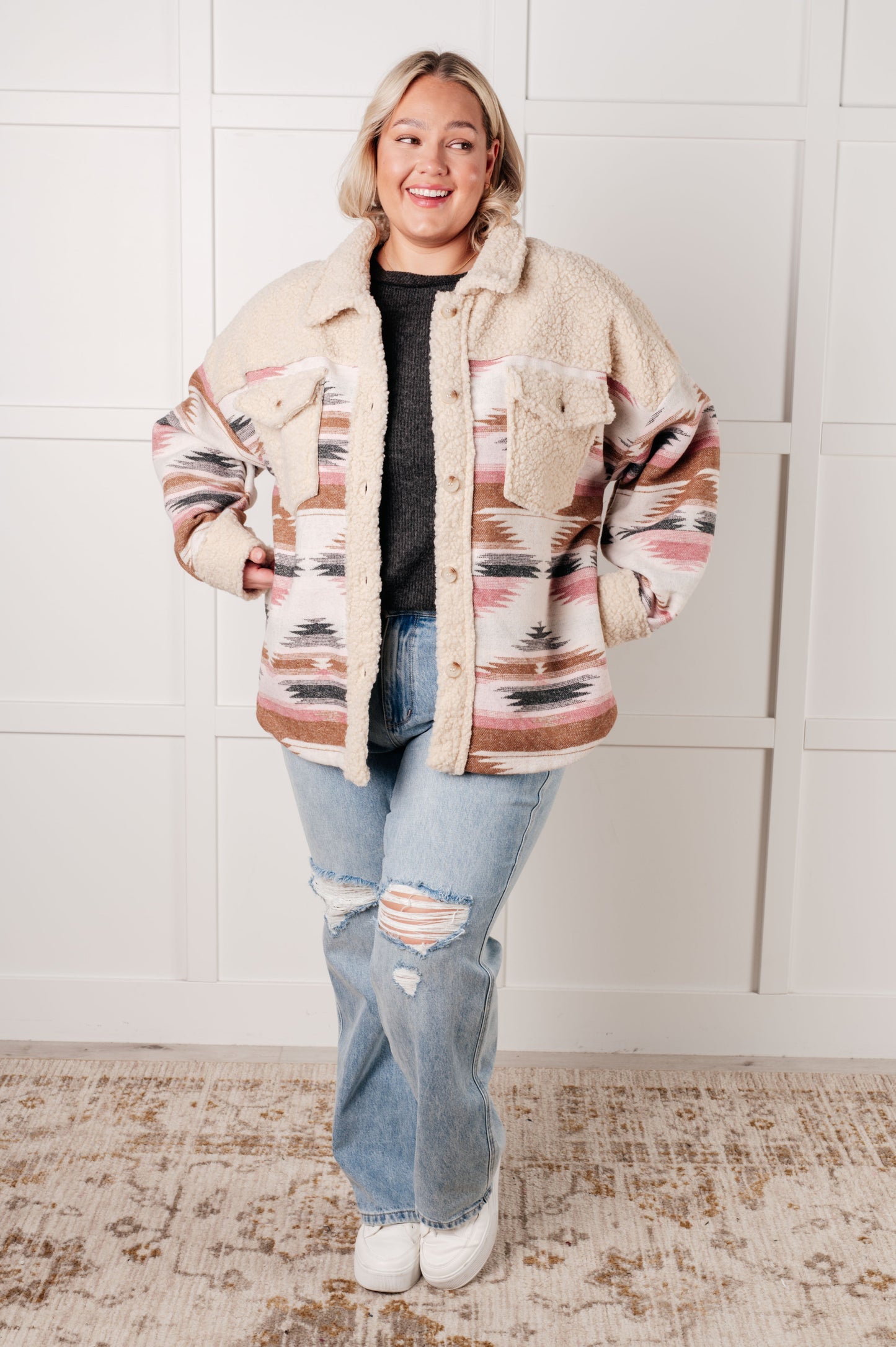 Person wearing a patterned jacket and jeans standing against a white tiled wall.