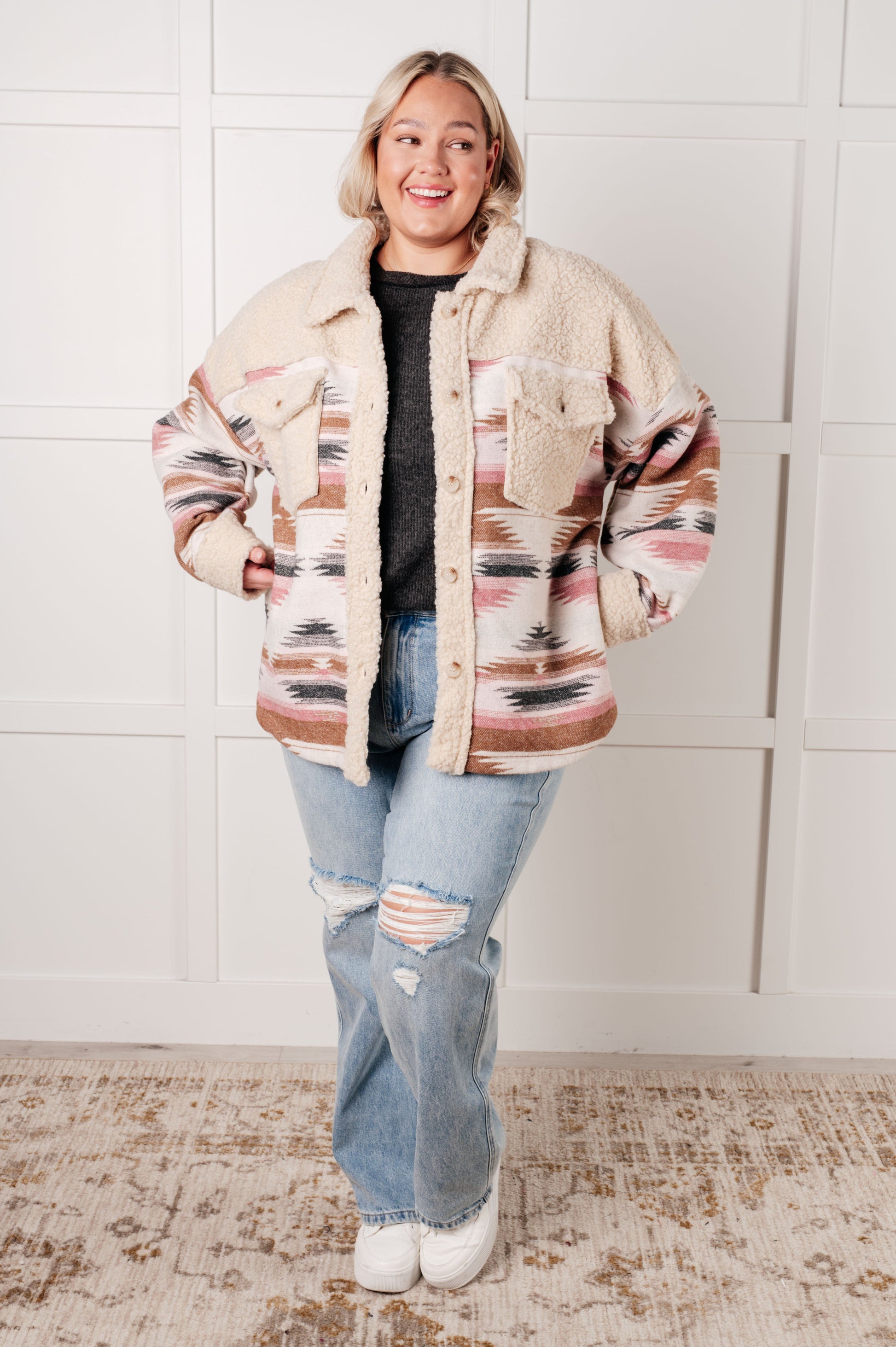 Person wearing a patterned jacket and jeans standing against a white tiled wall.