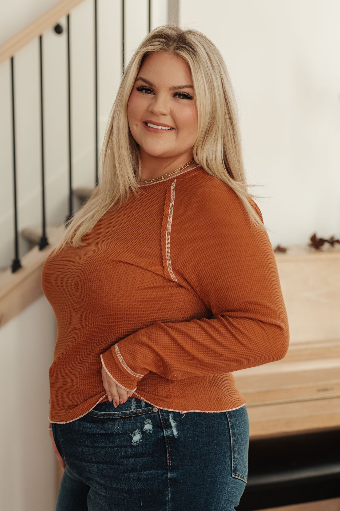 Woman wearing a rust-colored sweater and blue jeans standing indoors.
