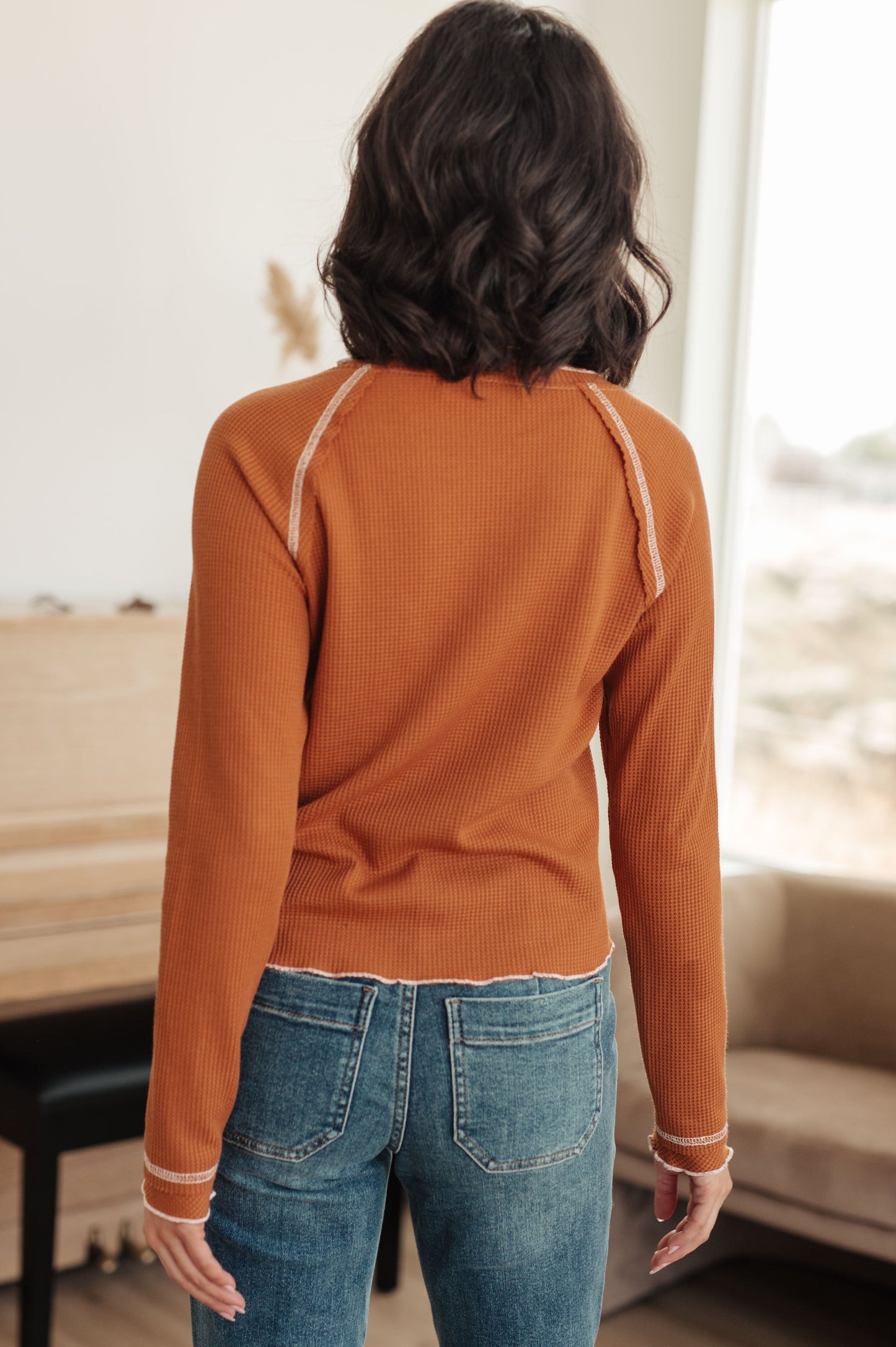 Person wearing an orange long-sleeve top and blue jeans in a room with a piano.