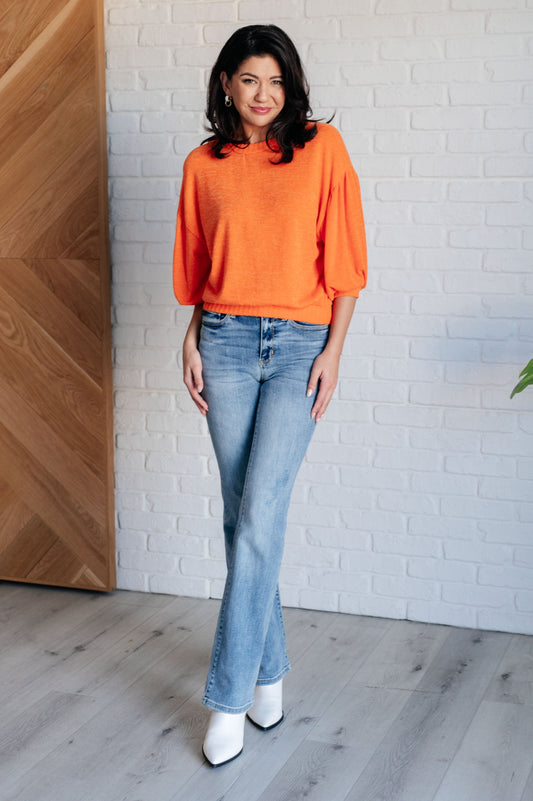 Woman wearing an orange sweater and blue jeans standing against a white brick wall.