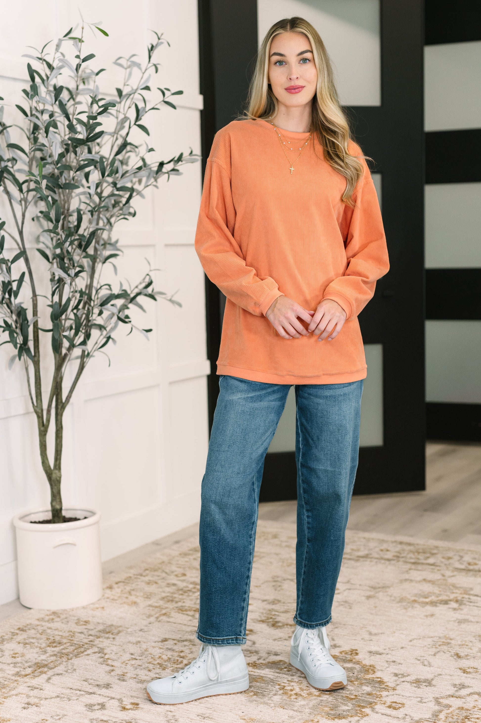 Woman wearing an orange sweater and blue jeans standing indoors with a plant and black and white striped wall in the background.