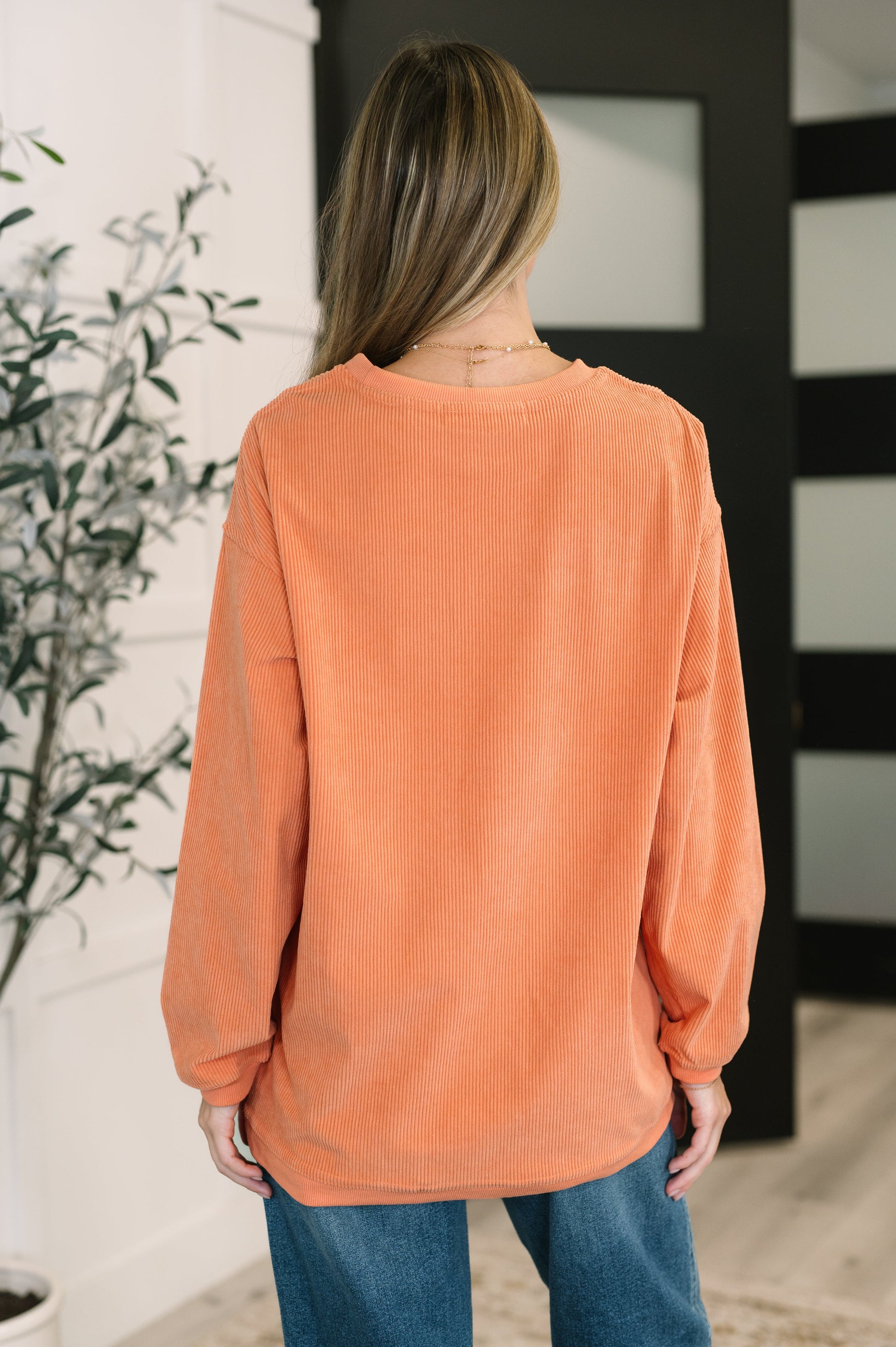 Person wearing an orange sweater and blue jeans in a room with a plant and mirror.