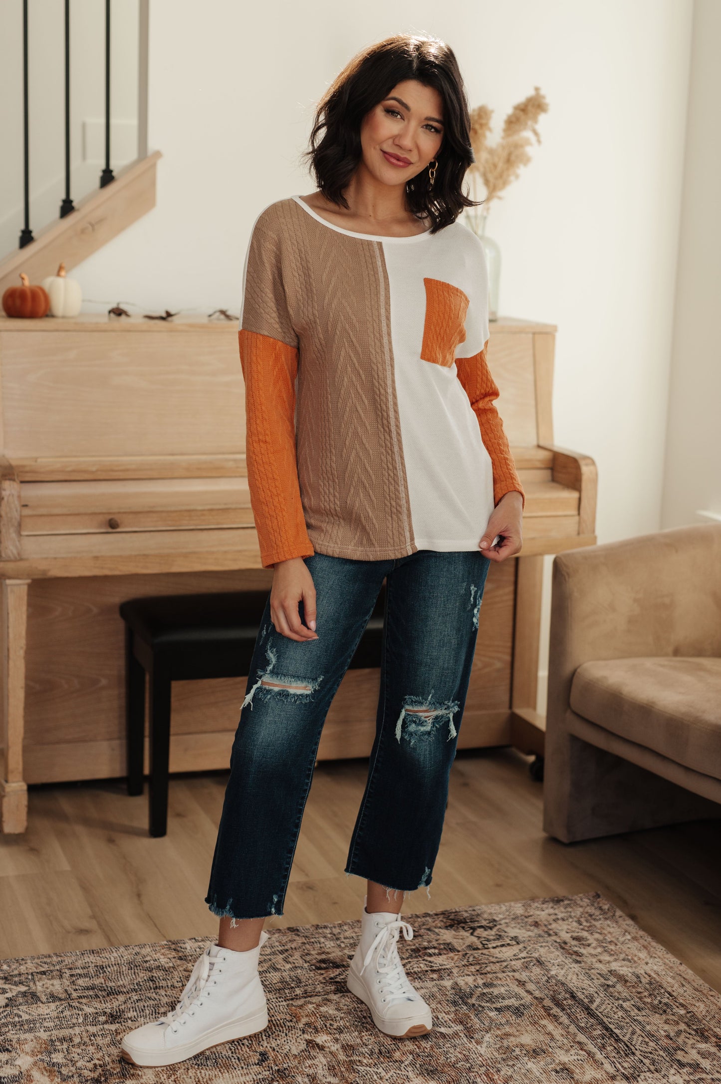 Woman wearing a color-blocked sweater and jeans standing in a room with a piano and sofa.