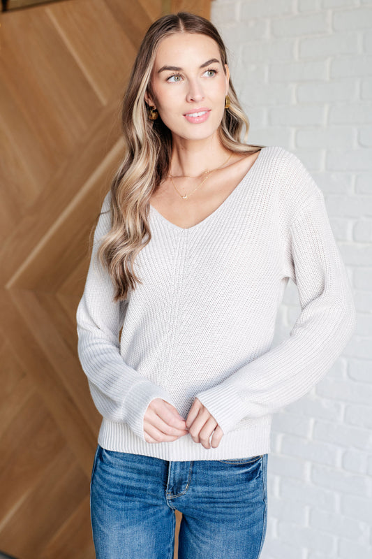 Woman wearing a light gray sweater and blue jeans against a wooden and white brick wall.