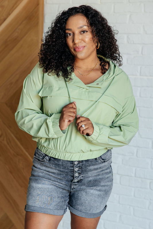 A woman wearing a light sage green hooded windbreaker jacket and gray denim shorts, standing against a split background of wood paneling and a white brick wall.