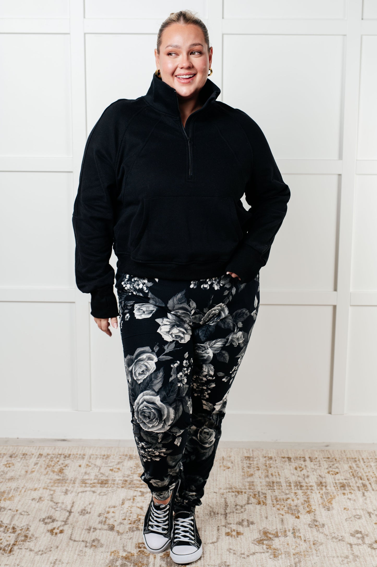 Person wearing a black jacket and floral pants standing against a white paneled wall.
