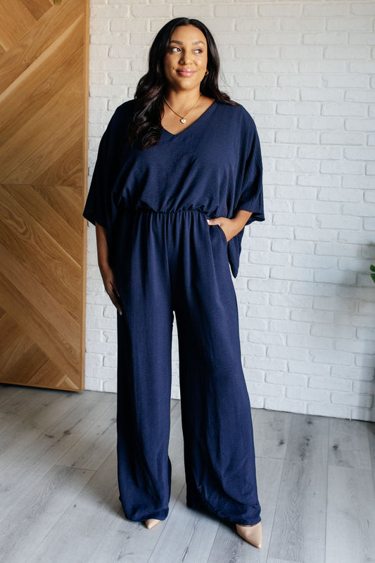 Woman wearing a navy blue jumpsuit standing against a white brick wall.