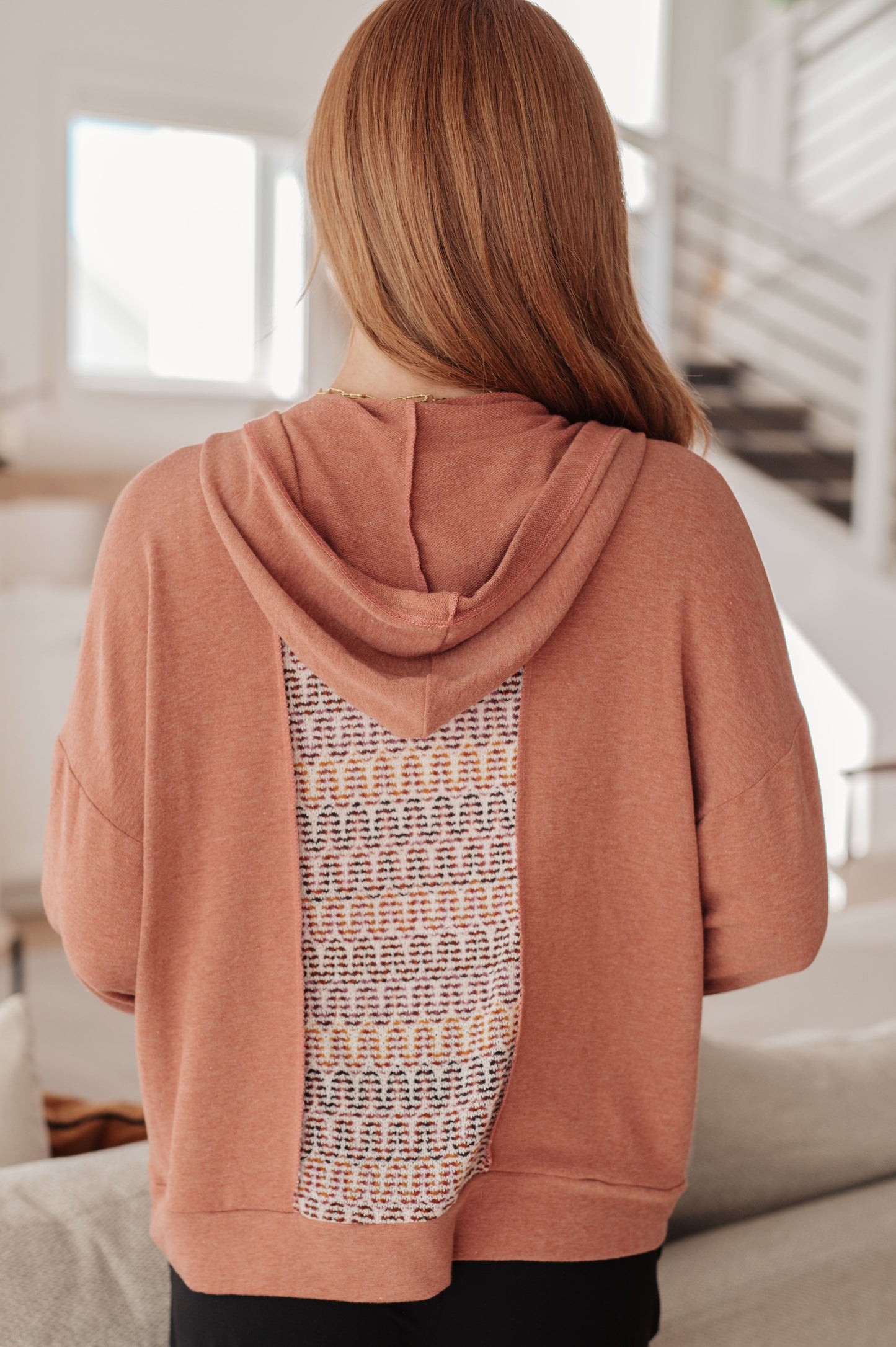 Person wearing a pink hoodie with a patterned back in a bright room.