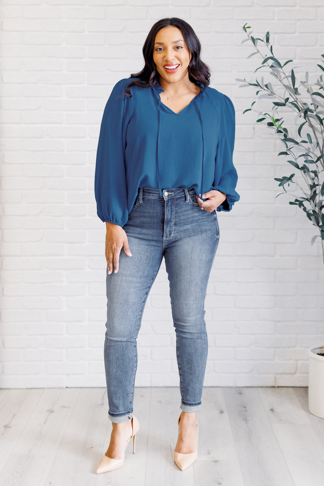 Woman wearing a blue blouse and jeans standing indoors with a white wall and plant in the background