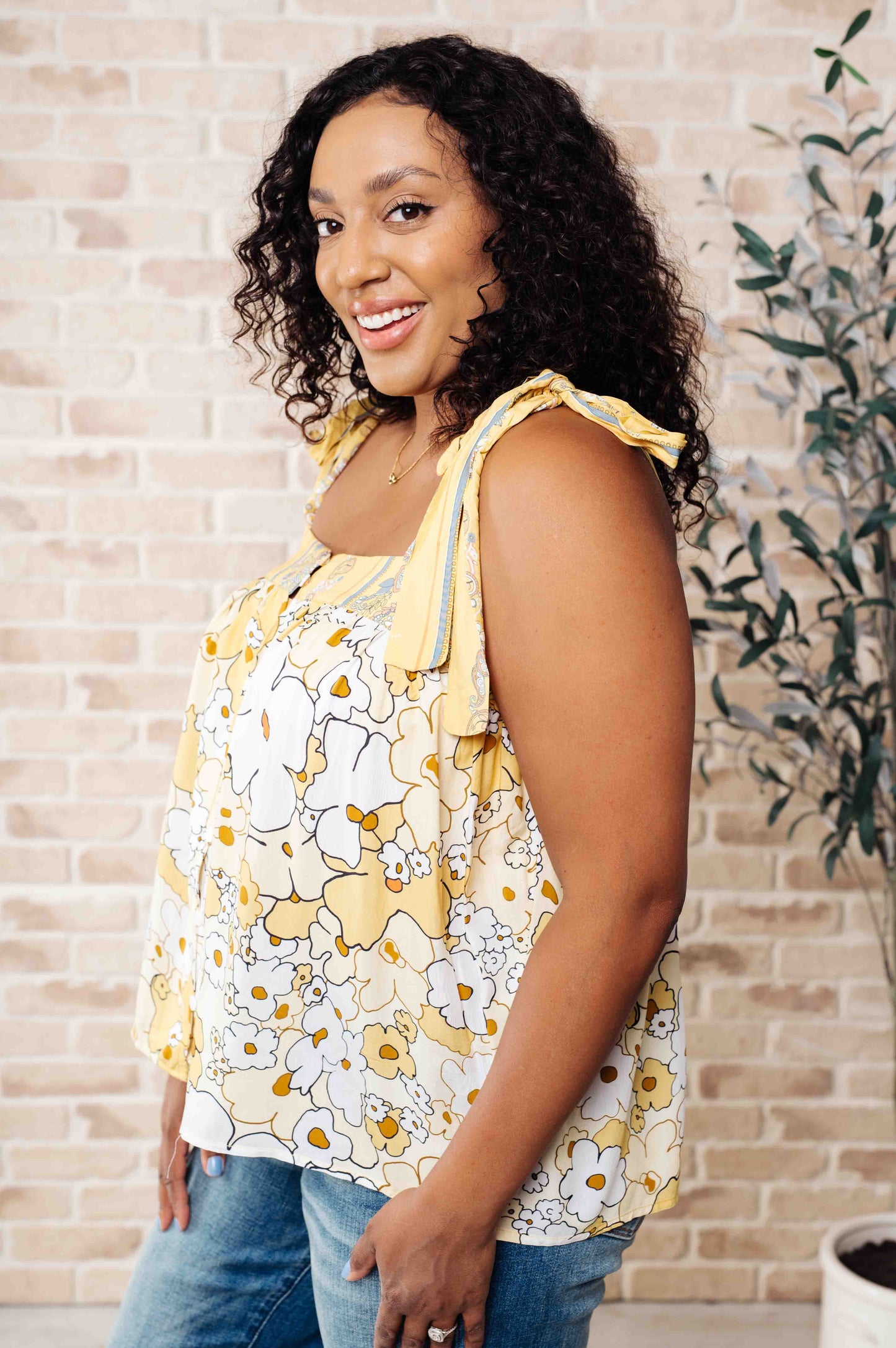 A woman wearing a floral tank top with yellow shoulder ties and a variety of jewelry, standing against a brick wall background.