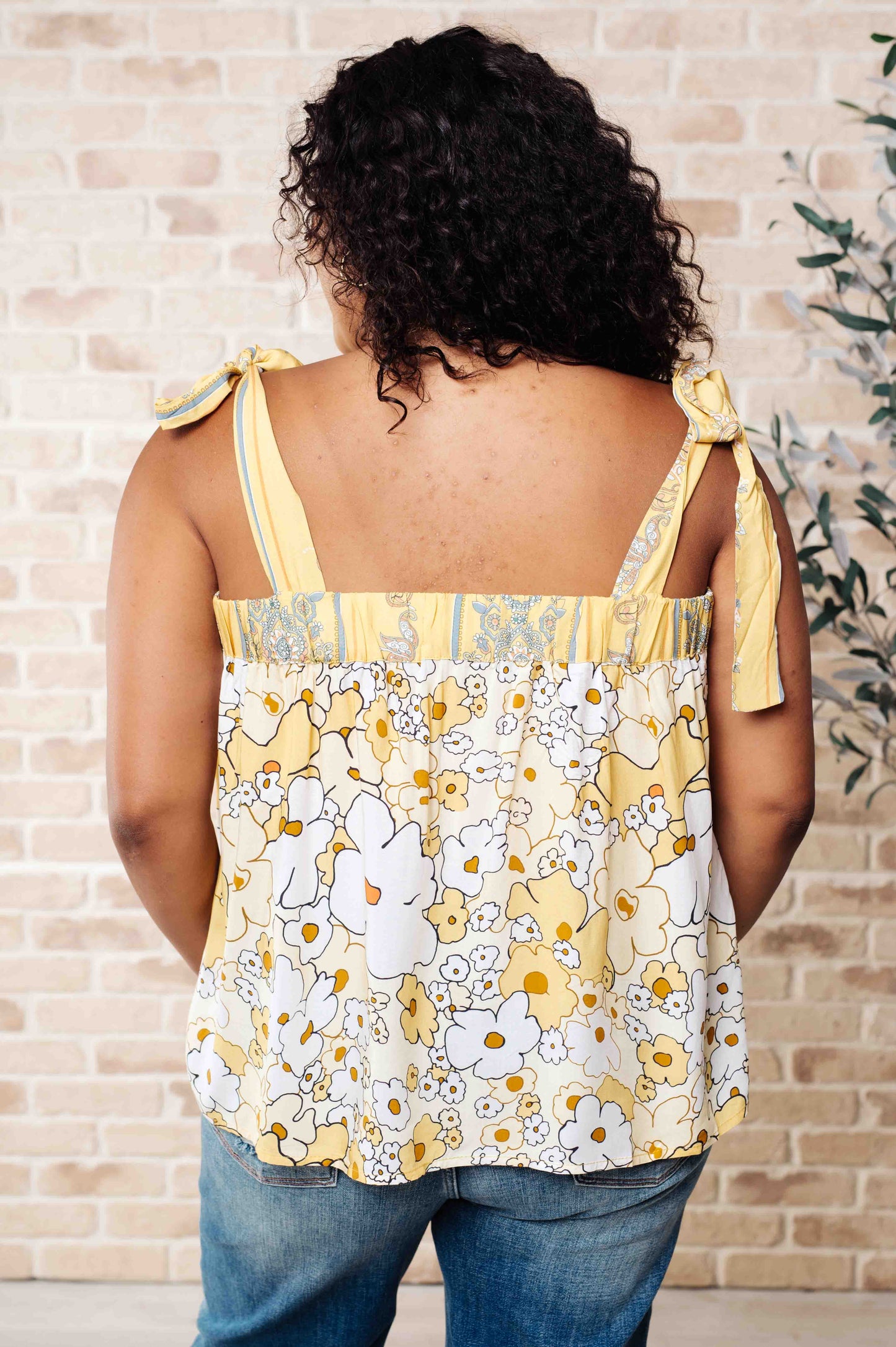 A woman wearing a floral tank top with yellow shoulder ties and a variety of jewelry, standing against a brick wall background.