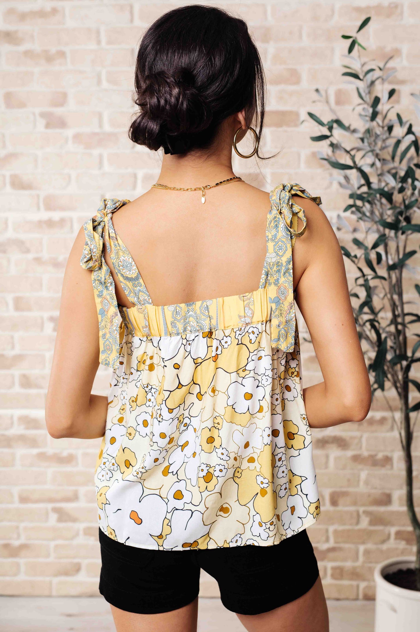 A woman wearing a floral tank top with yellow shoulder ties and a variety of jewelry, standing against a brick wall background.