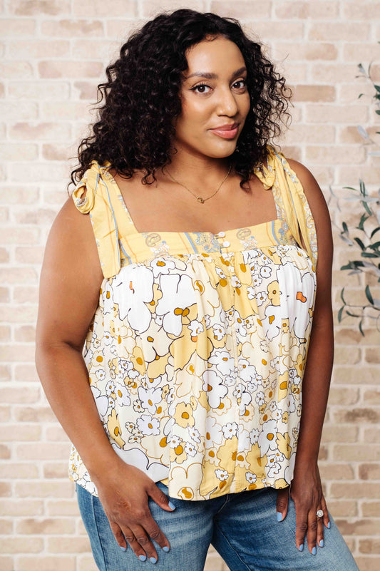A woman wearing a floral tank top with yellow shoulder ties and a variety of jewelry, standing against a brick wall background.