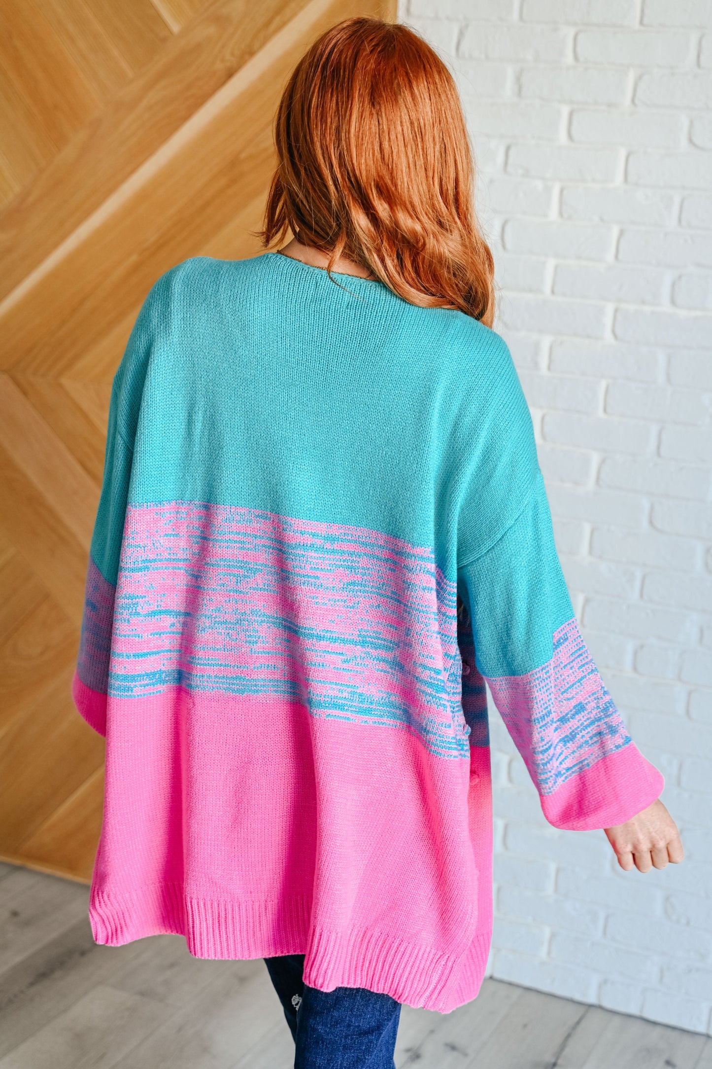 Person wearing a teal and pink sweater with a white brick wall background