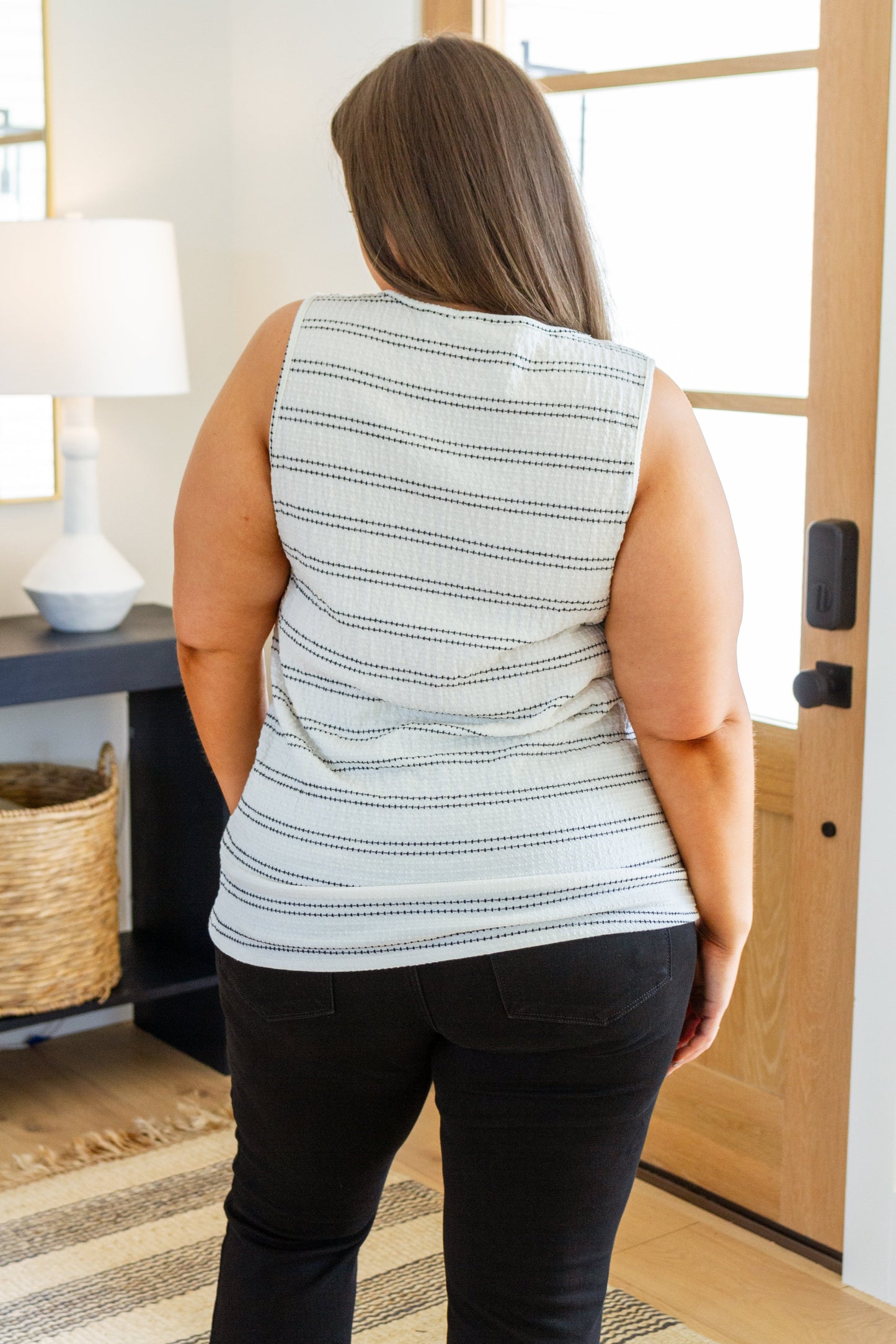 Person wearing a sleeveless patterned top and black pants in a room with a lamp and basket.