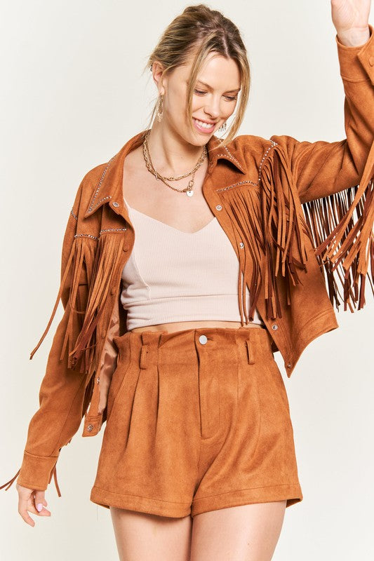 Woman wearing a brown suede jacket with fringe, beige top, and shorts on a light background